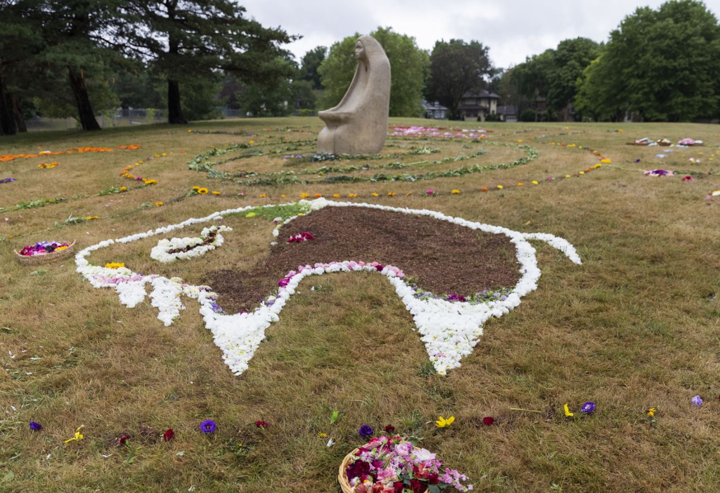 A buffalo drawn in flowers on the ground of a park, with a stone statue of a woman in the background. 