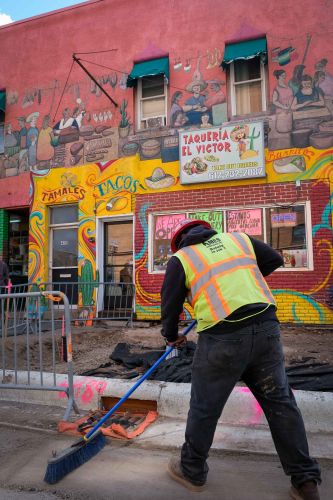 man wearing construction gear sweeps brick and ruble in front of building
