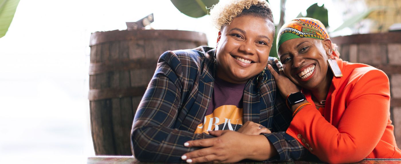 Drs. Brittany and Joi Lewis sitting at a table, and smiling while leaning on each other. 
