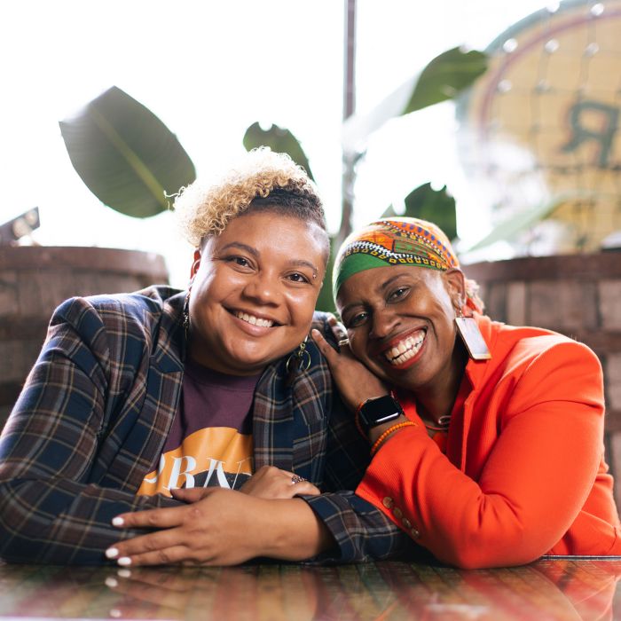 Drs. Brittany and Joi Lewis sitting at a table, and smiling while leaning on each other. 