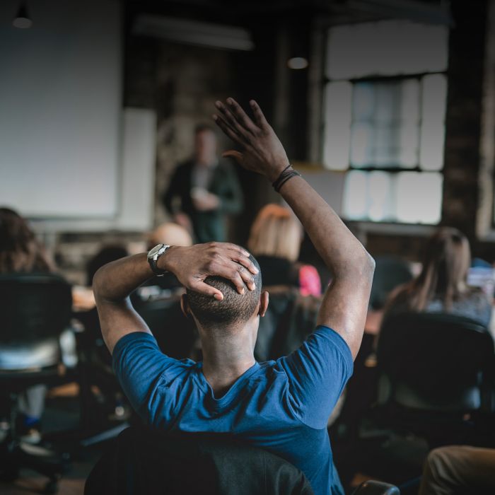 Man, shown seated from behind, raises hand to speak in an information session